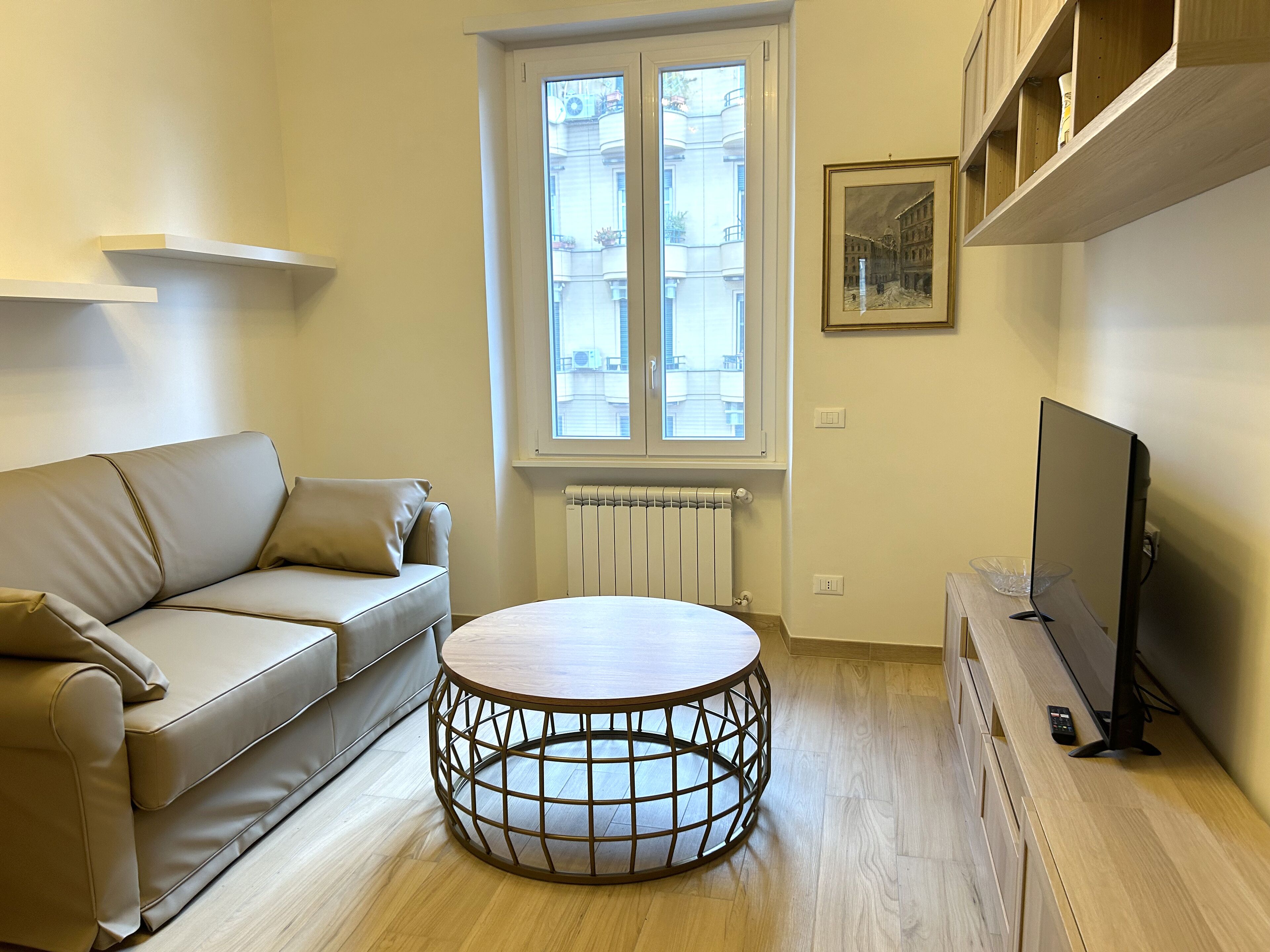 A modern living room with a beige sofa, a round coffee table, a television, and a window with a view of buildings.
