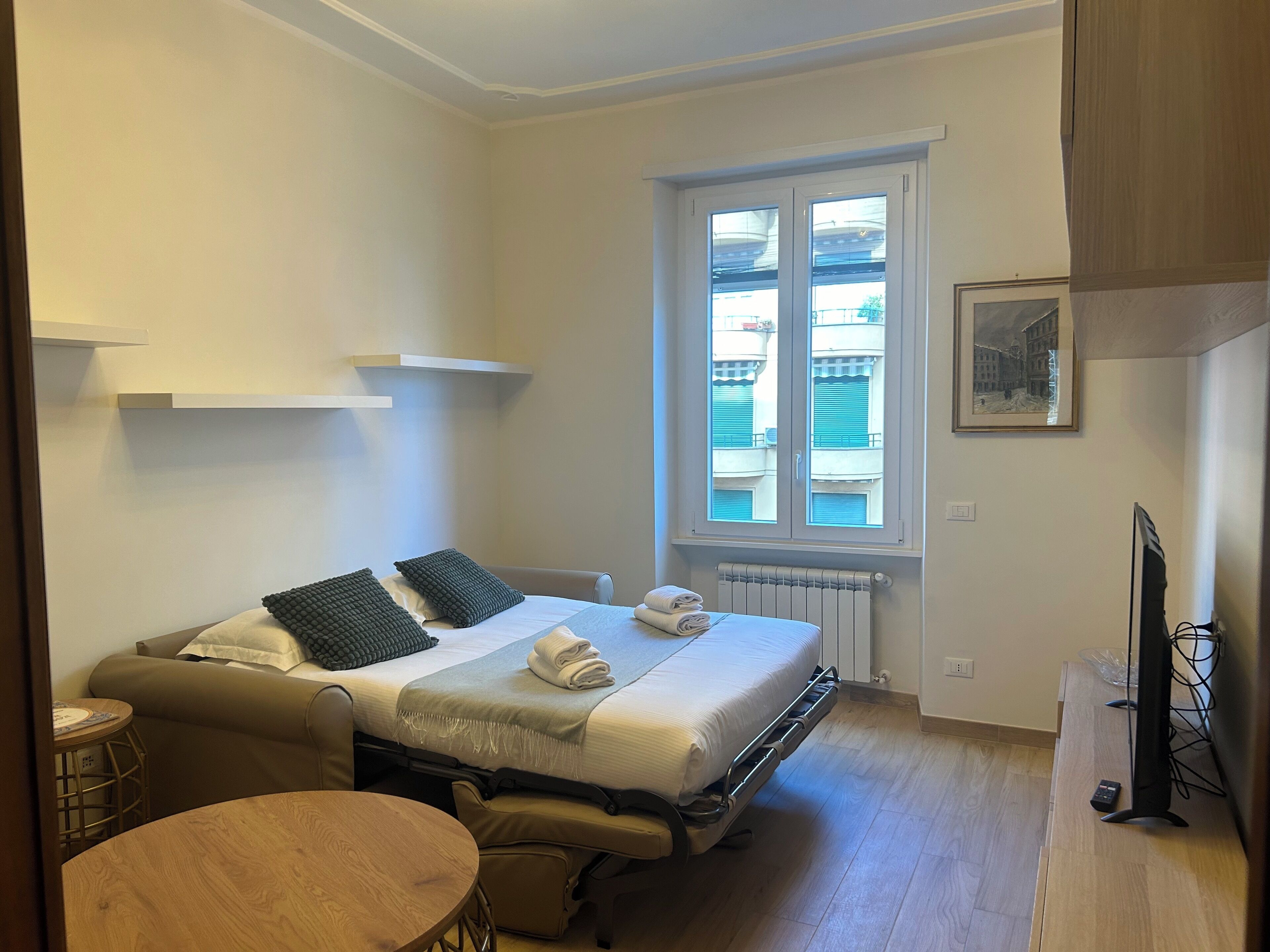 A compact hotel room with a bed, a small table, a sofa, and a window with a view of buildings.