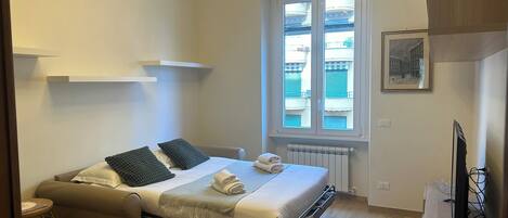 A compact hotel room with a bed, a small table, a sofa, and a window with a view of buildings.