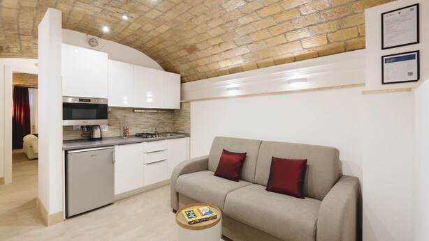Una cocina moderna con armarios blancos, horno empotrado y microondas. Hay un sofá color beige con cojines rojos y un taburete de madera con un libro. El techo muestra ladrillo visto.