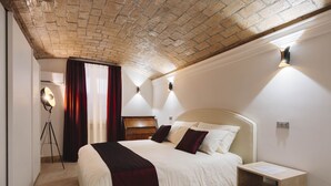Habitación de hotel con techo de ladrillo, cama con ropa de cama blanca y rojo oscuro, mesita de noche con lámpara y un pequeño escritorio con lámpara.