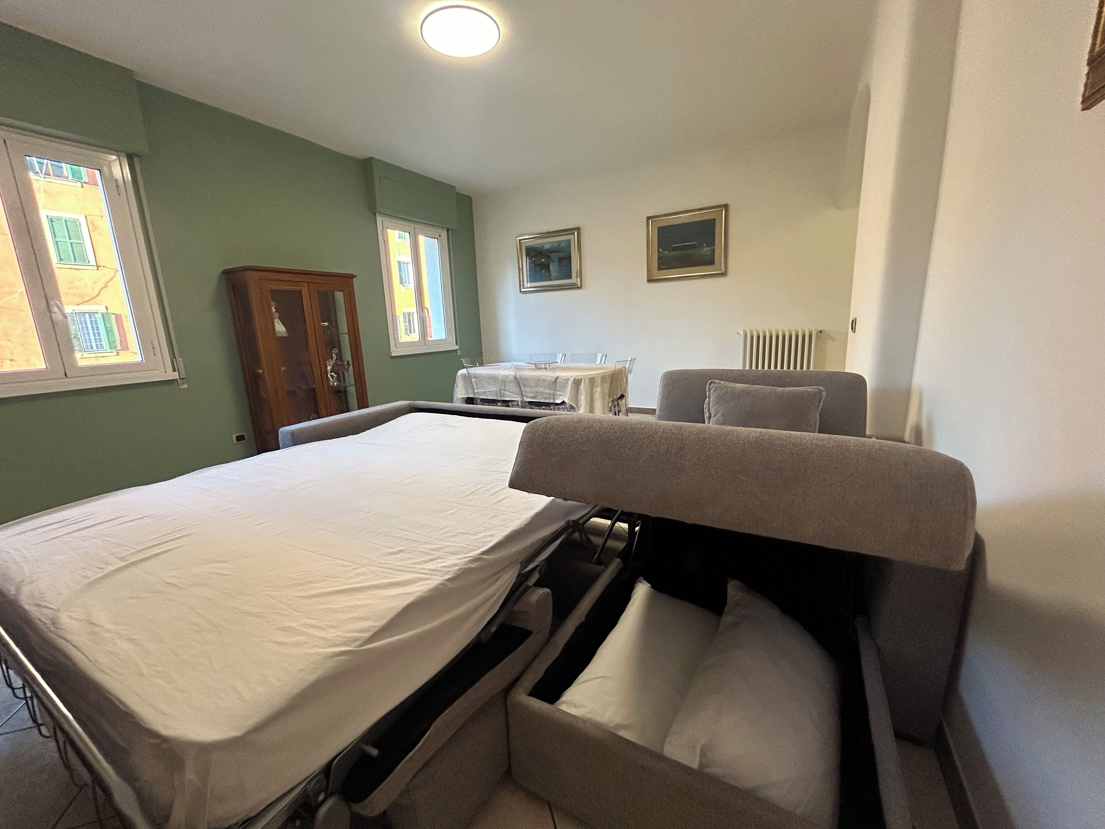 Habitación con cama individual y sofá cama, ventana con persianas, radiador y una pequeña mesa con una silla.