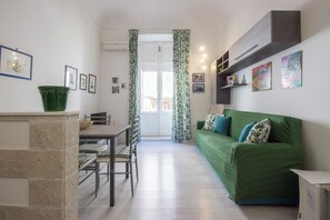 A compact living space with a dining area, a green sofa, and a kitchen counter. - Maniace Homes - Angelica by Ortigia Apartments (Syracuse)