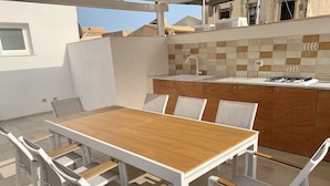 An outdoor patio with a wooden table and white chairs, a tiled backsplash, and a small kitchen area with a gas stove. - Amate Case by Ortigia Apartments (Syracuse)