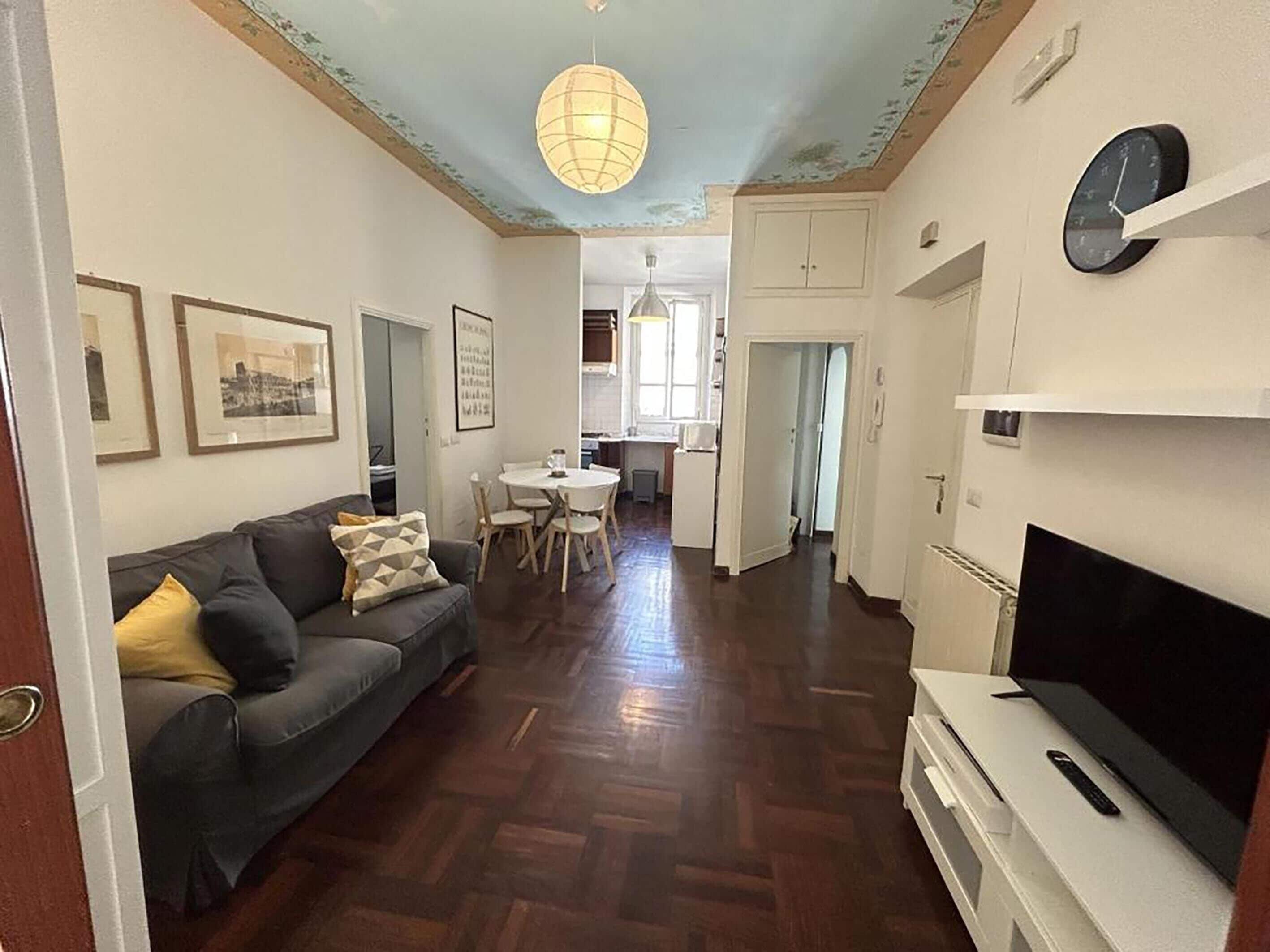 A living room with a sofa, a dining table with chairs, a kitchen area, and a TV on a stand.
