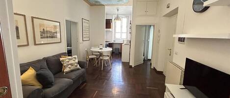 A living room with a sofa, a dining table with chairs, a kitchen area, and a TV on a stand.
