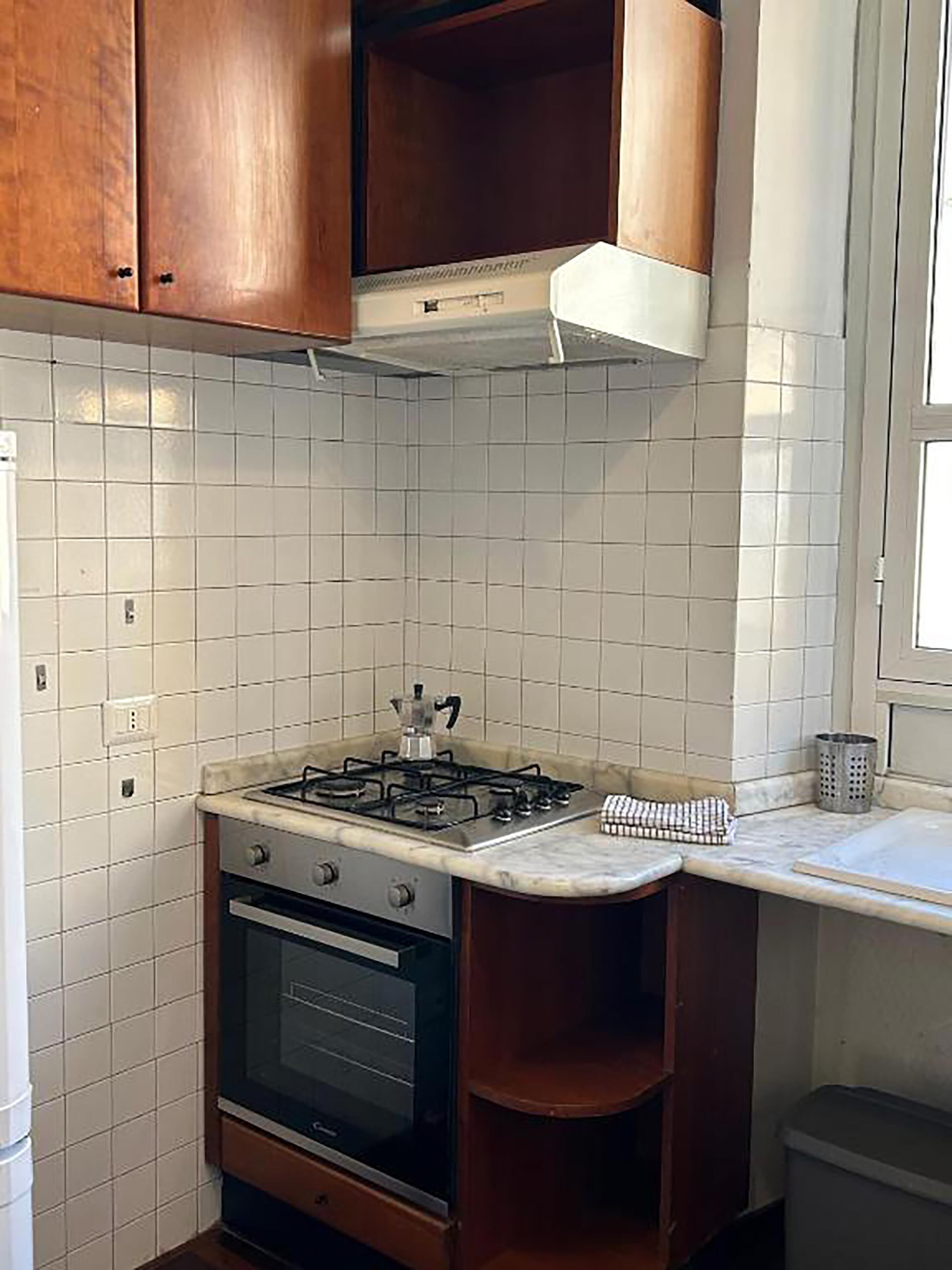 Een keuken met witte betegelde muren, een gasfornuis, een oven, houten keukenkastjes en een raam.