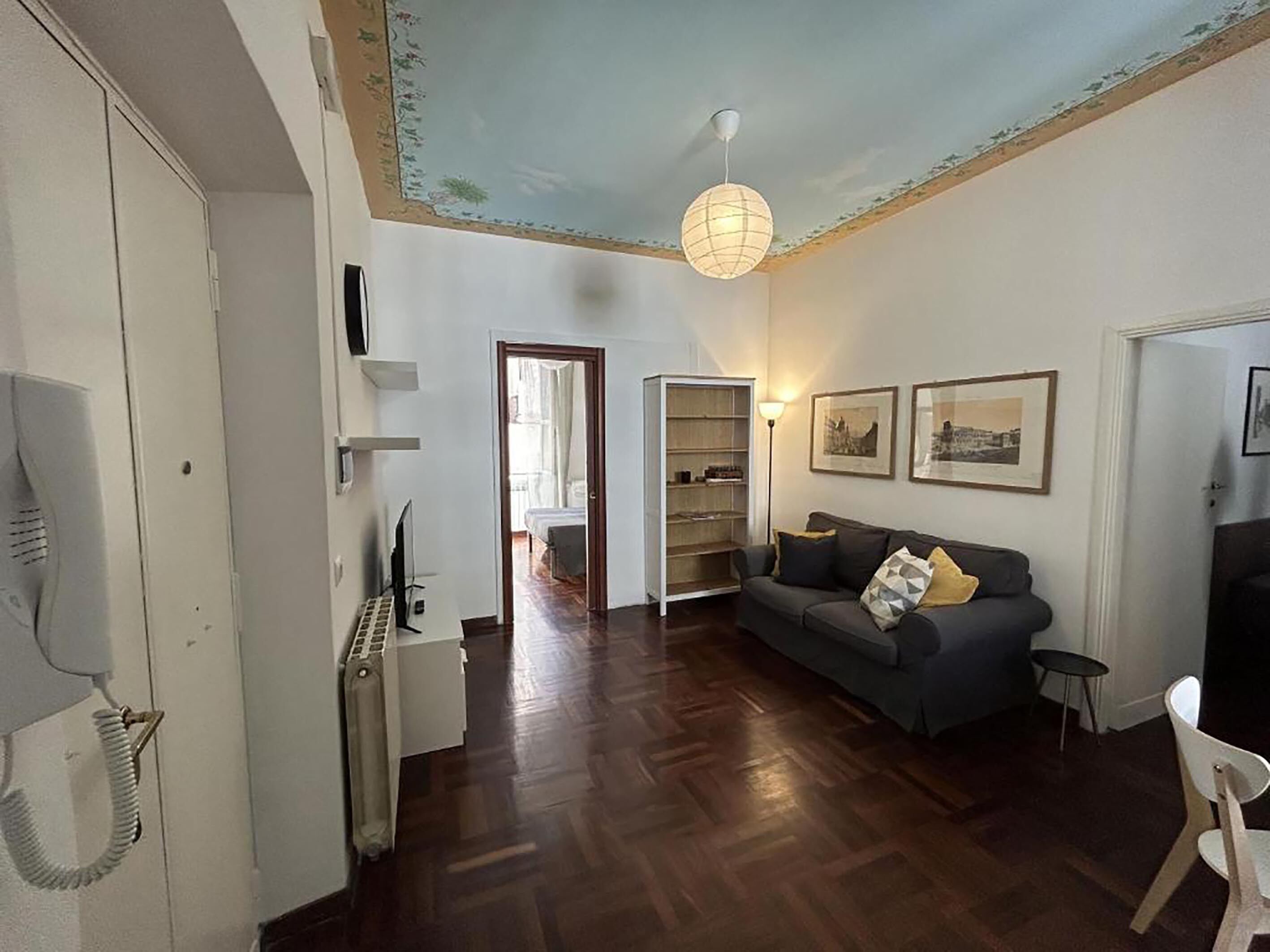 A living room with a dark wooden floor, a grey sofa with cushions, a white radiator, a ceiling light fixture, and a door leading to another room.
