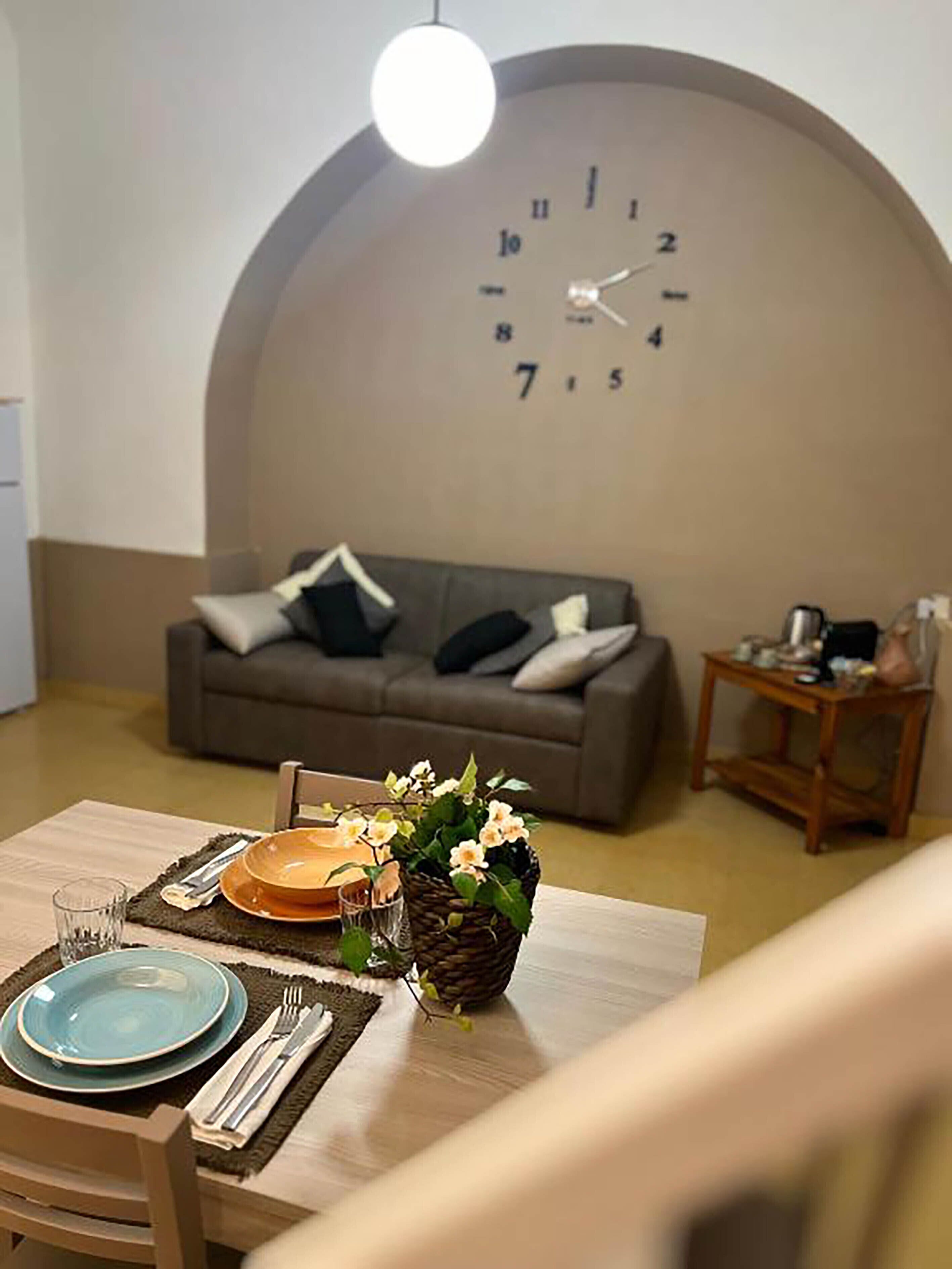 A dining area with a table set for two, a clock on the wall, and a sofa in the background.