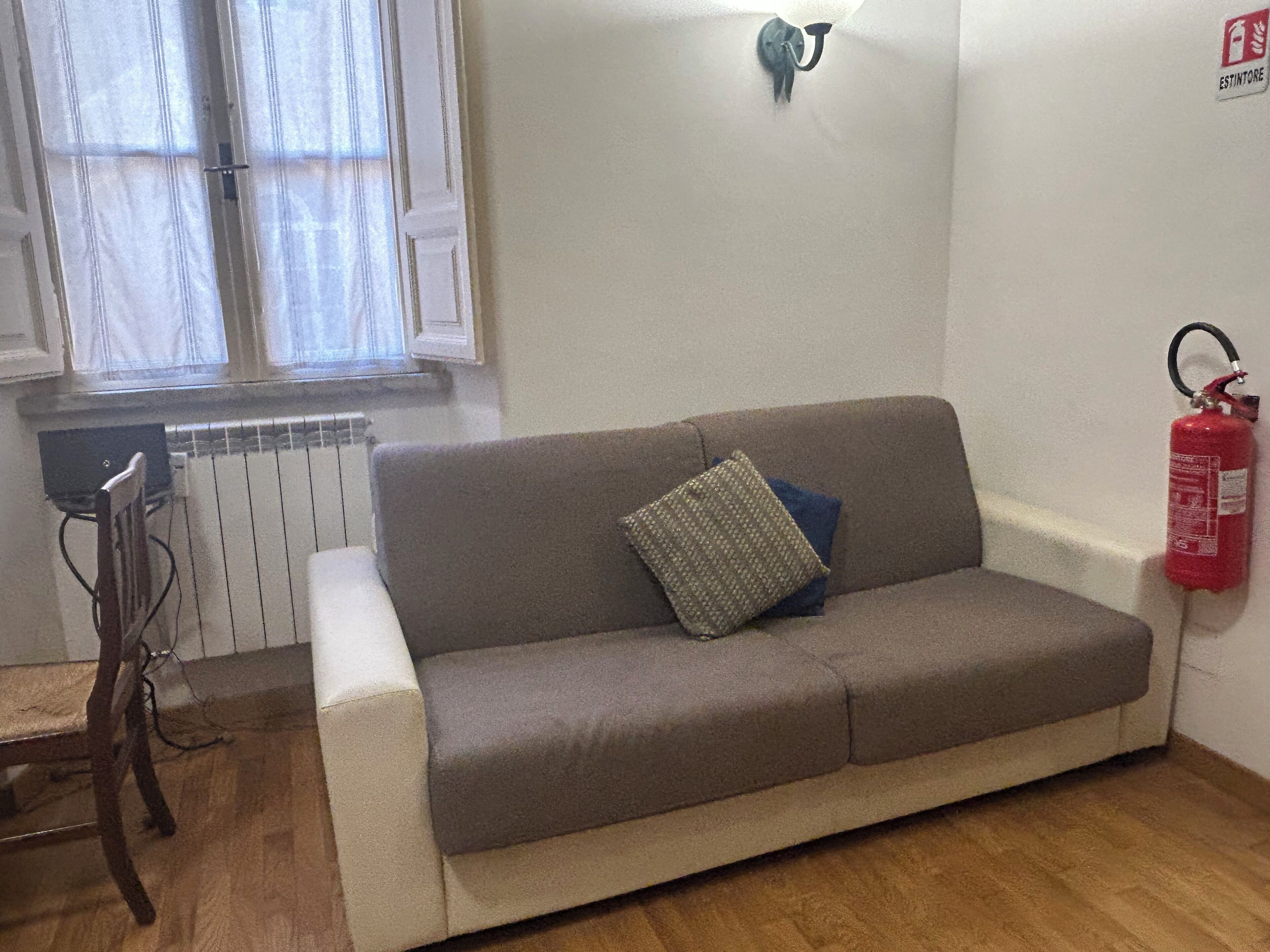 Ein Zimmer mit einer Couch, einem Heizkörper, einem Fenster mit Vorhängen, einer Wandleuchte und einem Feuerlöscher.