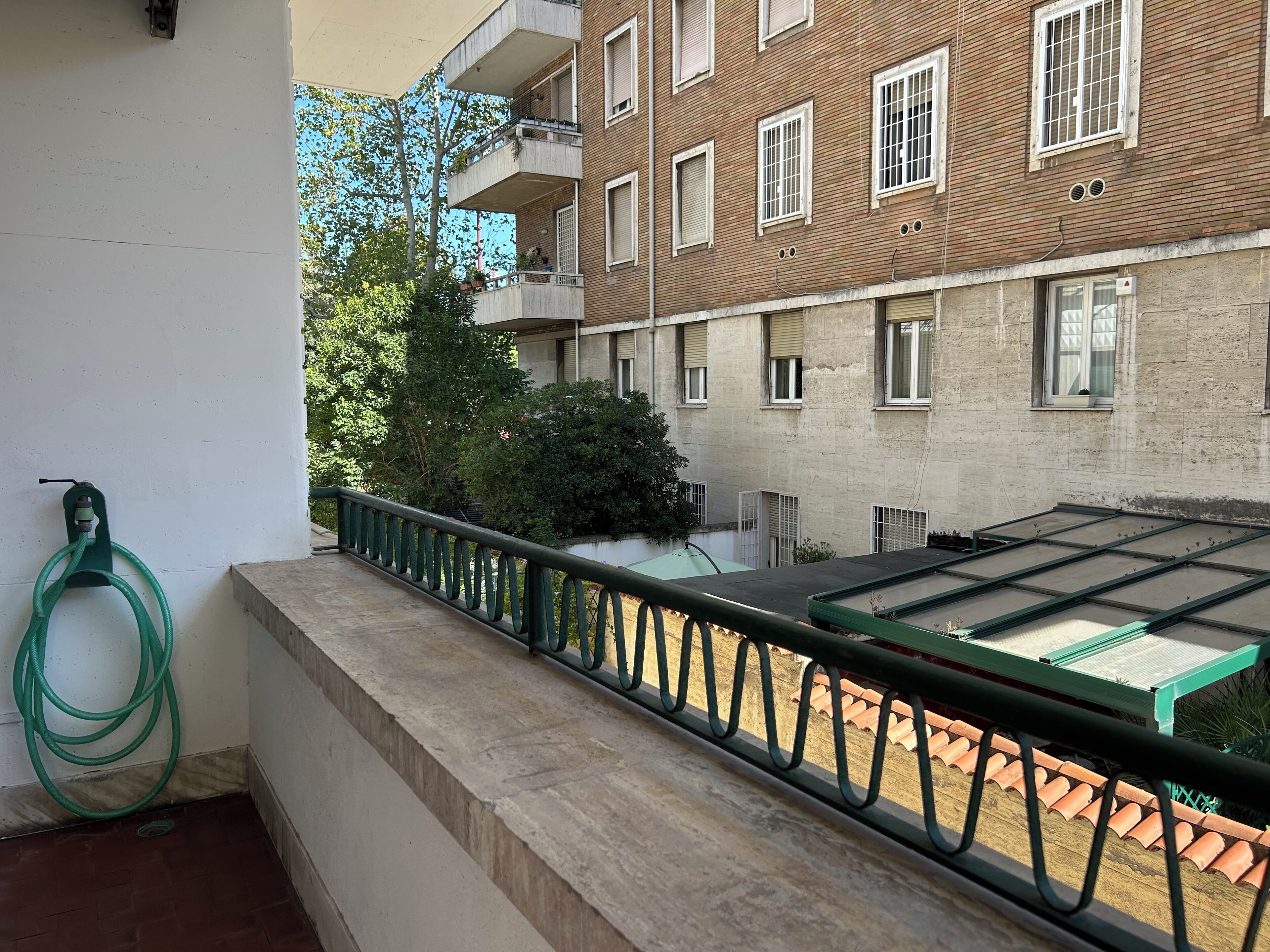A balcony with a green hose, a metal railing, and a view of a brick building with multiple windows and balconies.