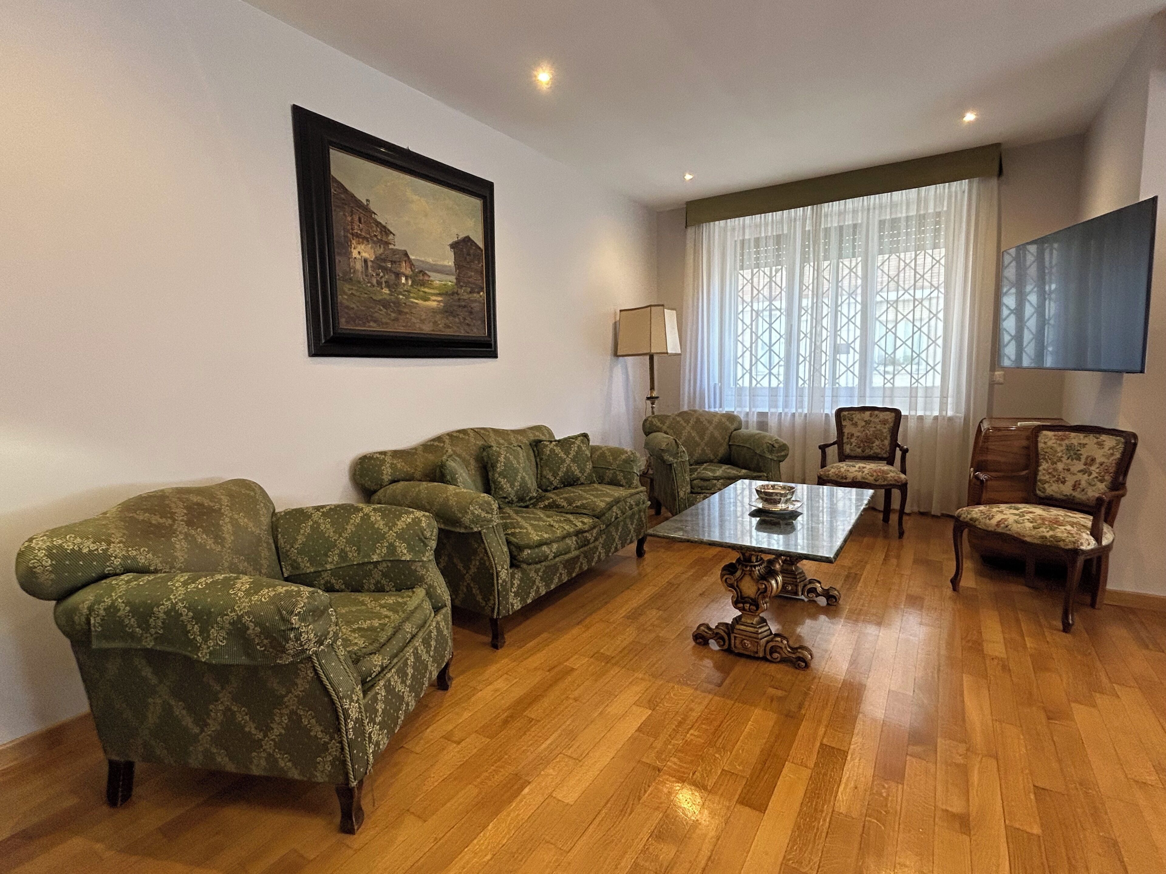 A living room with a wooden floor, green patterned sofas, a coffee table, and a painting on the wall.