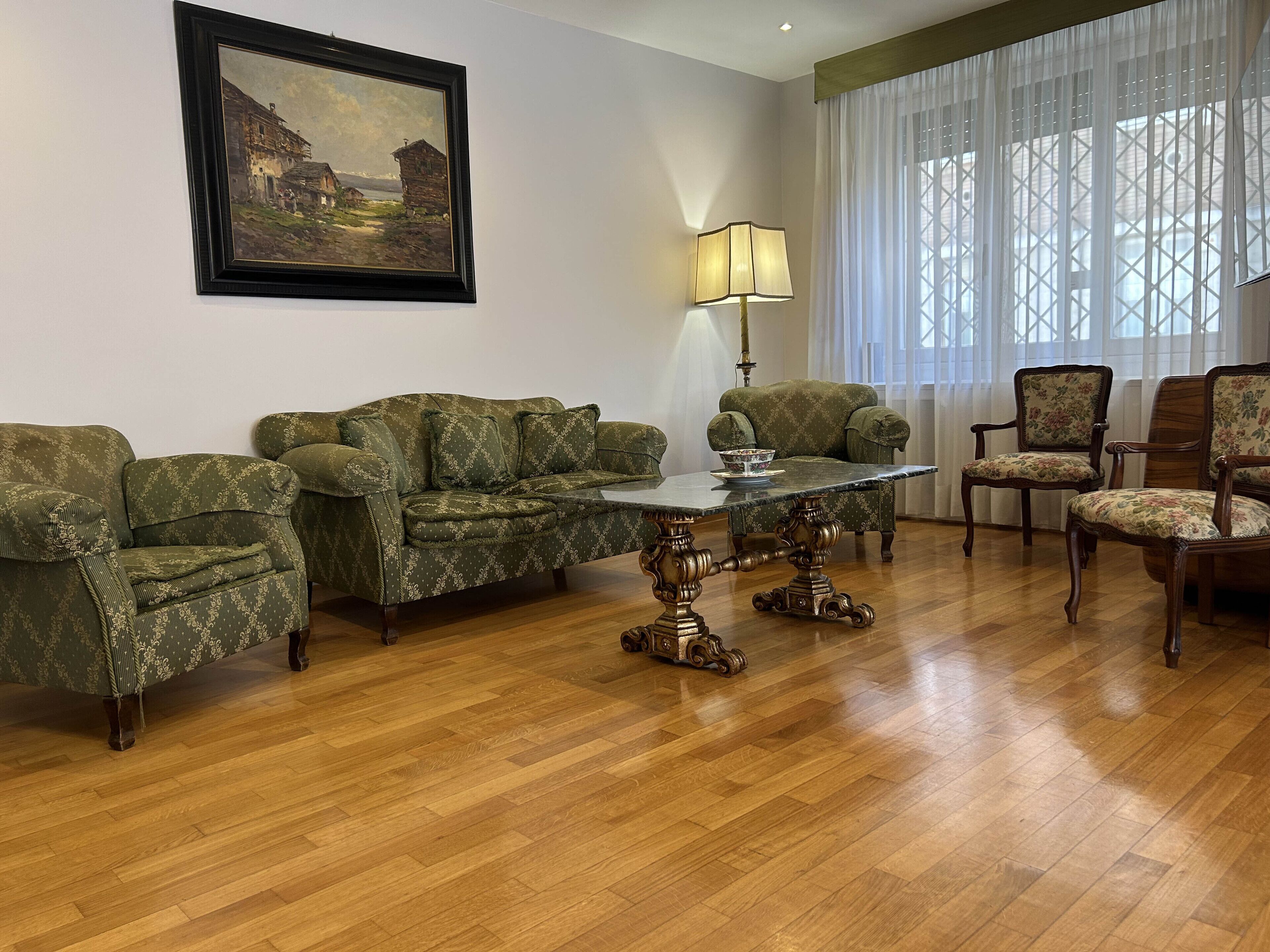 A living room with a wooden floor, green patterned sofas, a glass coffee table, and a painting on the wall.