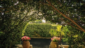 Um caminho serpenteia por um jardim exuberante, com um sebe ao fundo e dois vasos grandes de flores cor-de-rosa de cada lado.
