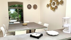 Uma área de jantar com uma mesa comprida, cadeiras brancas e uma máquina de café. Há pratos decorativos na parede e uma tigela sobre a mesa.