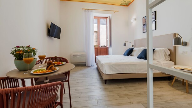 Une chambre d’hôtel de taille réduite comprenant un lit, un coin repas avec une table et des chaises, un téléviseur fixé au mur et une fenêtre avec des rideaux.