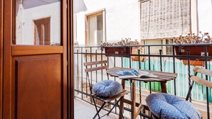 Un balcon avec une table en bois, deux chaises et une fenêtre avec un store blanc.