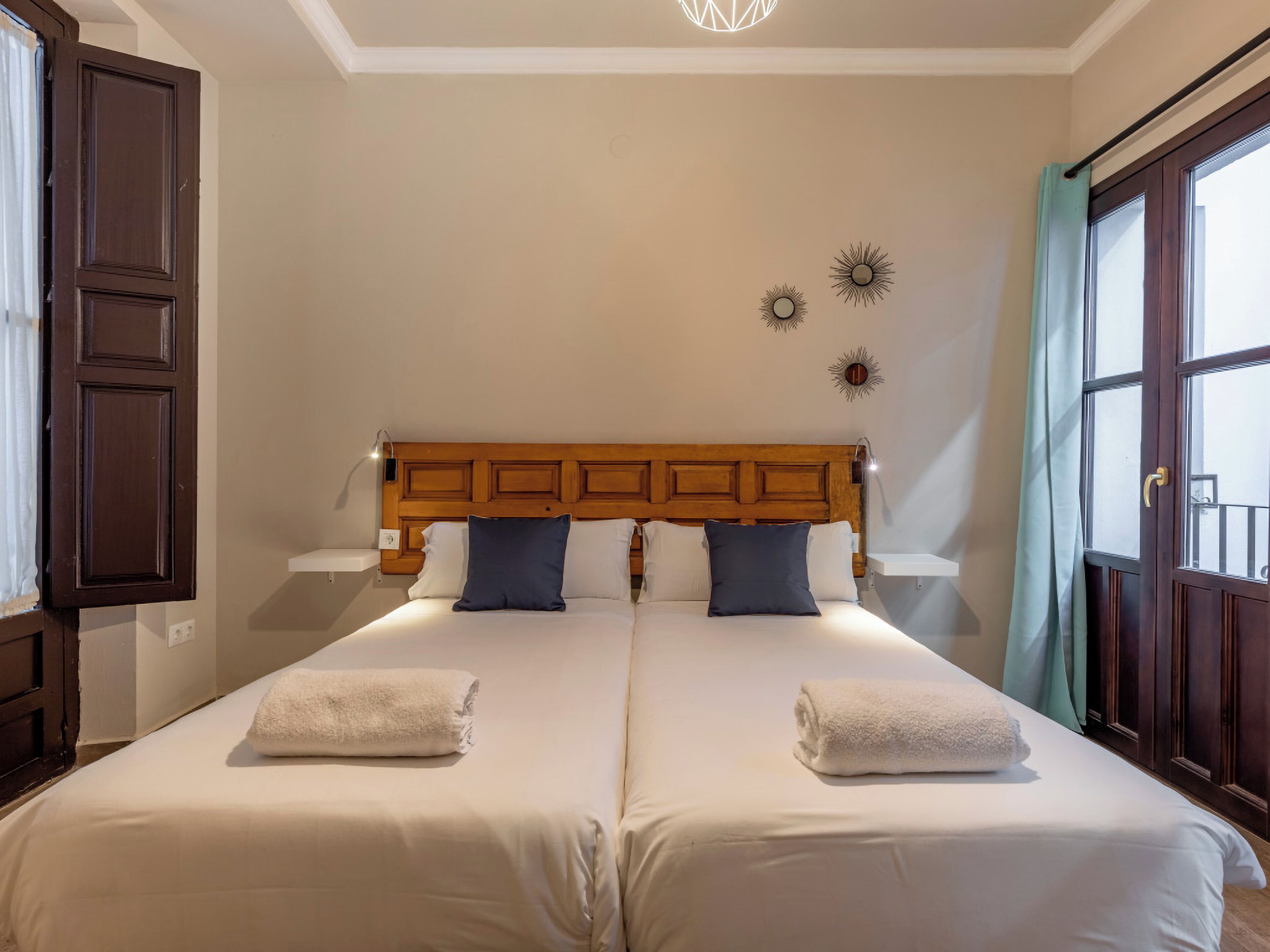 A double bed with two pillows and two towels, a wooden headboard, wall sconces, and a decorative sun motif.