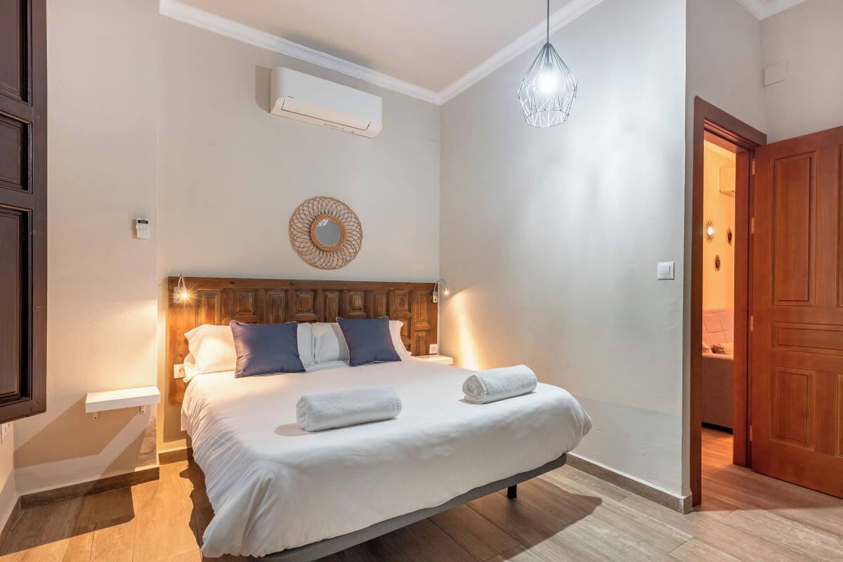 A bedroom with a bed, a wooden headboard, a wall-mounted air conditioner, a hanging light fixture, and a wooden door.
