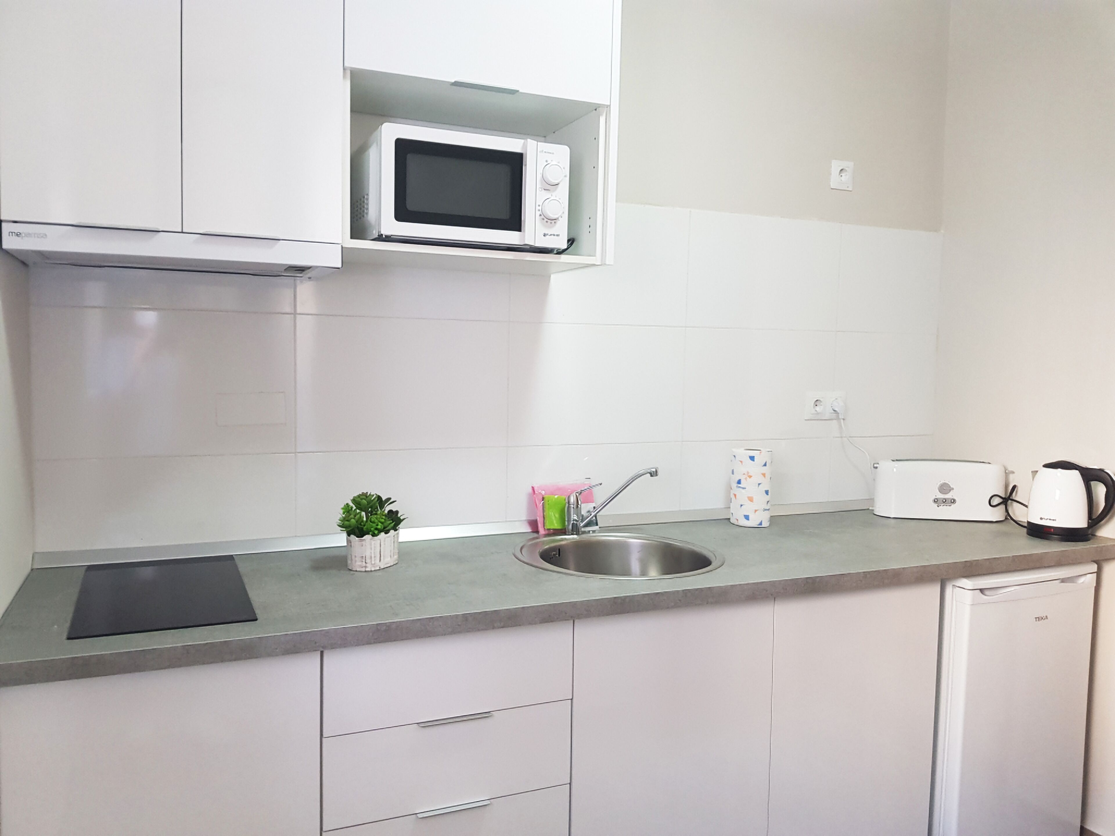Una cocina con gabinetes blancos, microondas, tostadora, hervidor, pileta y una pequeña planta en maceta.