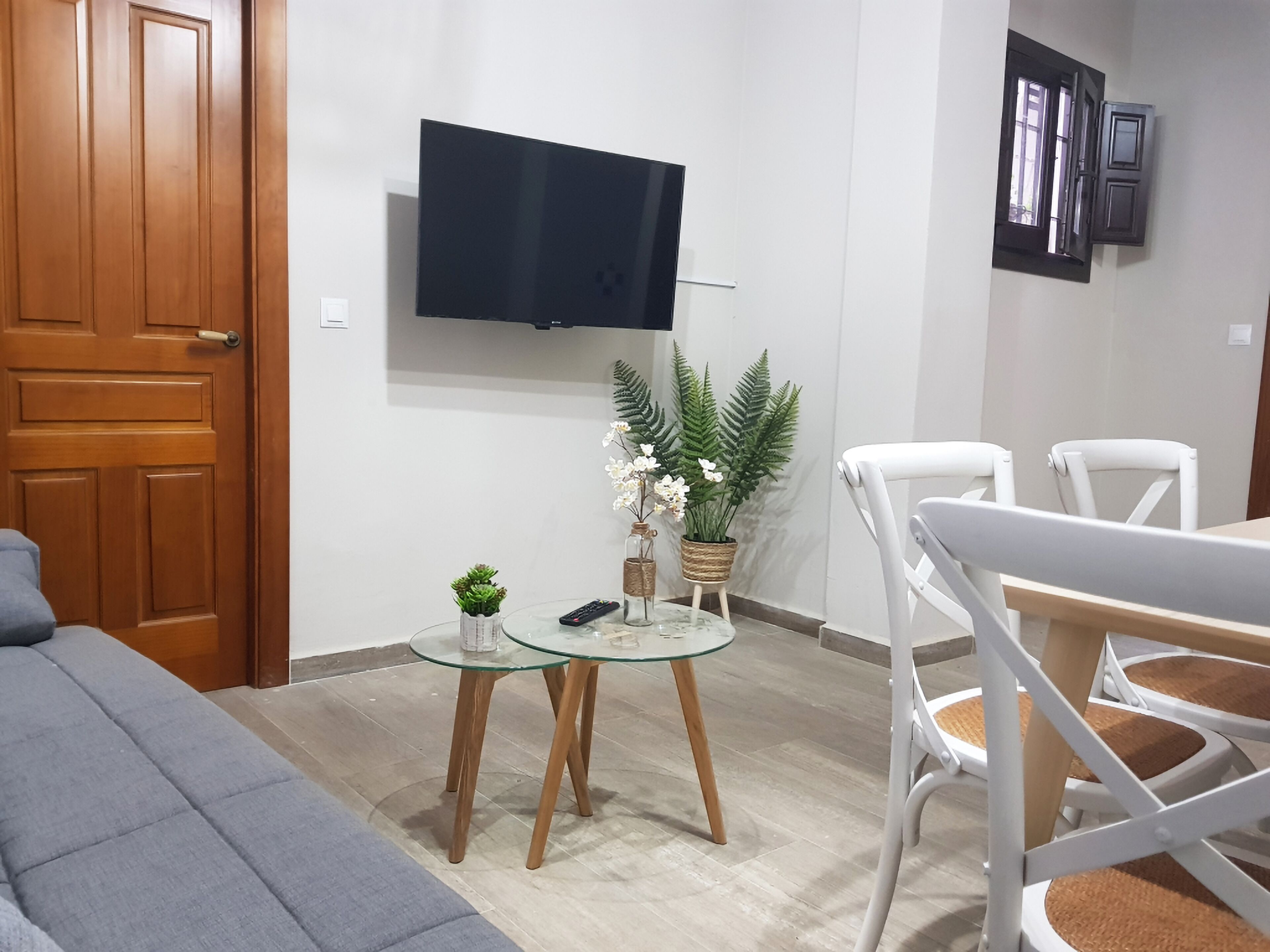 A living room with a grey sofa, a glass coffee table with decorative items, a white dining table with chairs, a mounted TV, and a wooden door.