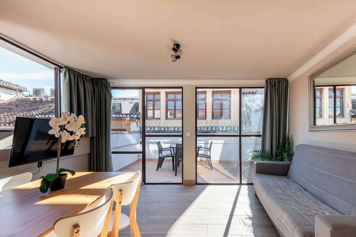 A modern living room with a sofa, a dining table with chairs, a TV, and a balcony with a view of buildings.