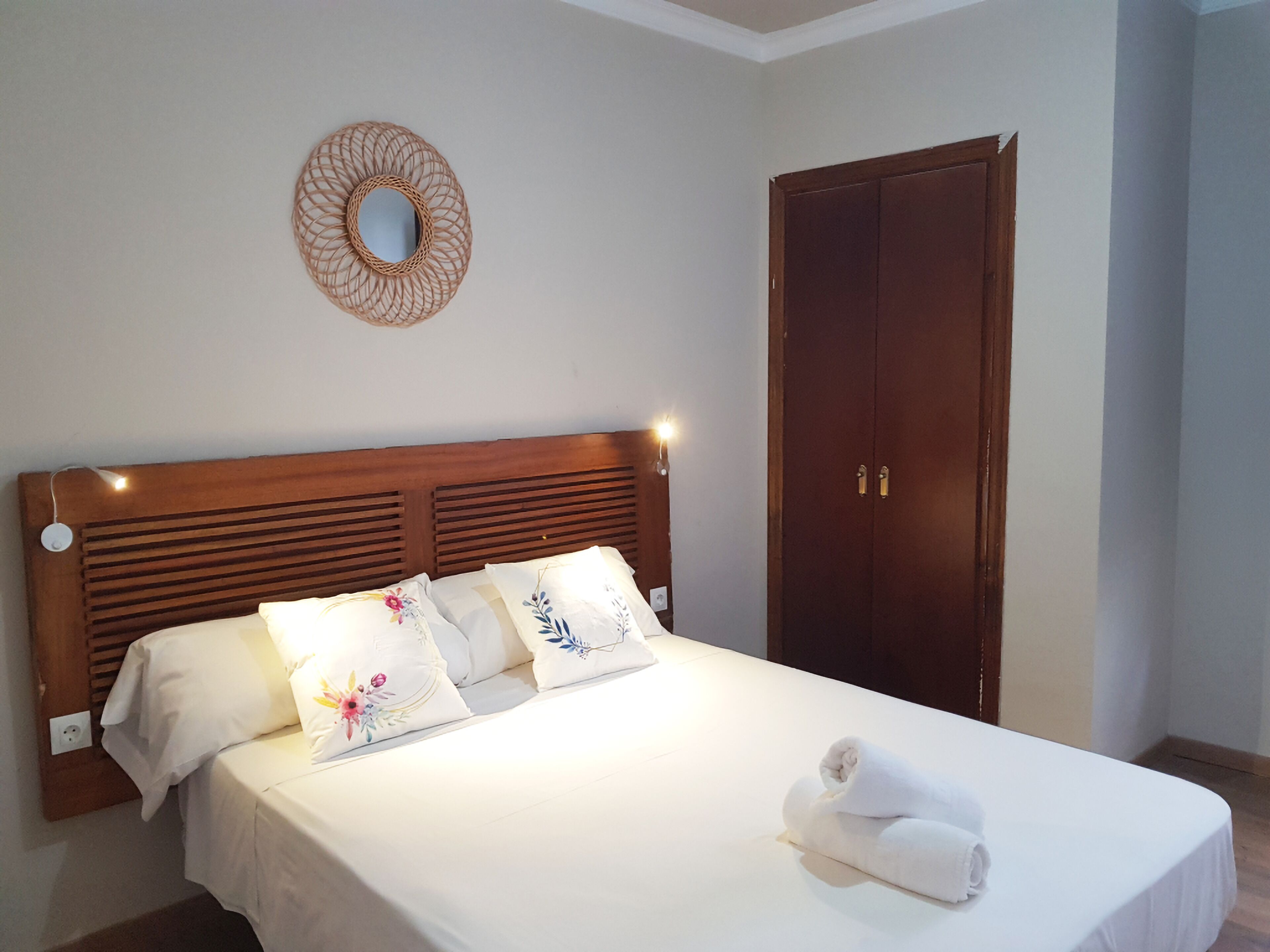 A neatly made bed with white linens and floral pillows, a wooden headboard, a round mirror on the wall, and a wooden wardrobe.
