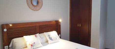 A neatly made bed with white linens and floral pillows, a wooden headboard, a round mirror on the wall, and a wooden wardrobe.
