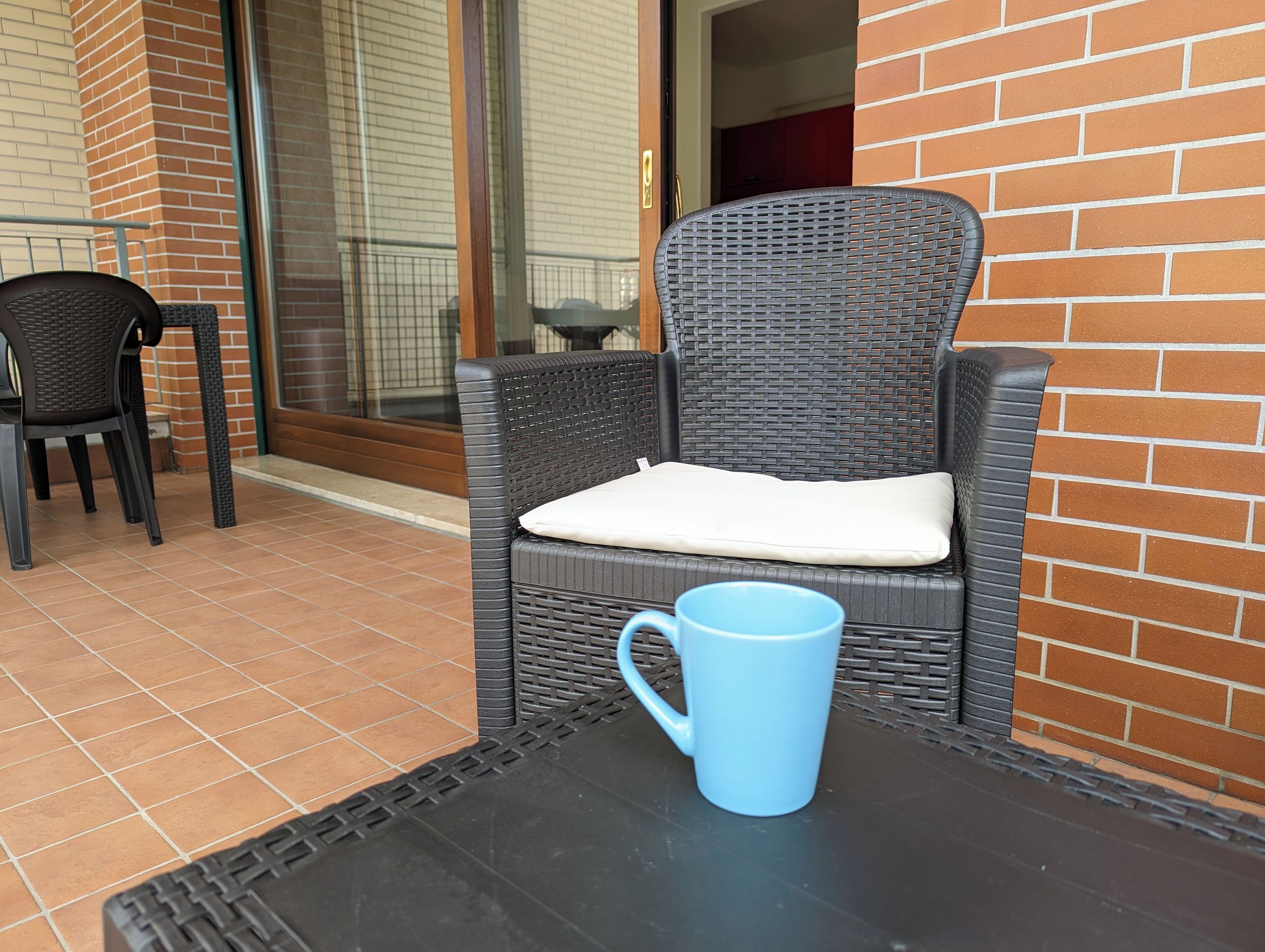 Una silla de mimbre con un cojín en un patio con baldosas, una taza azul sobre una mesa y un muro de ladrillo al fondo.