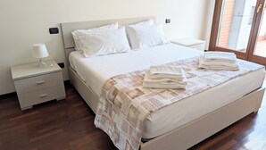 A neatly made bed with white linens and a checkered blanket, a bedside table with a lamp, and a view of a brick building through a window. - Spina Verde (Como)