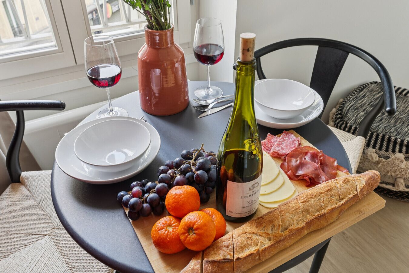 A table set with a bottle of wine, glasses, plates, a loaf of bread, grapes, and oranges.
