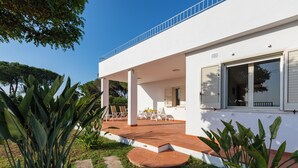 Una casa moderna con un porche cubierto, mobiliario exterior y abundante vegetación.