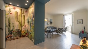A modern interior with a cactus-themed wall mural, a dining area with a gray sofa and patterned chairs, and a balcony with a railing. - Casa Lumia by Ortigia Apartments (Syracuse)