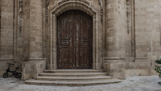 A stone building with a large wooden door, steps leading up to it, and a small bicycle parked nearby. - Casa Lumia by Ortigia Apartments (Syracuse)