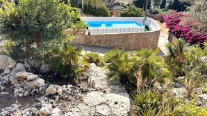 A pool surrounded by a stone wall, with various plants and a view of the sea.