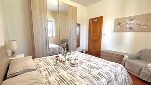 A neatly made bed with a patterned bedspread, a bedside table with a lamp, a chair, a door, and a wall-mounted artwork.