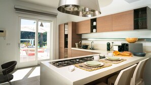 A modern kitchen with a central island, stainless steel appliances, and a large window overlooking a patio area. - Villa Elena by Ortigia Apartments (Isola)