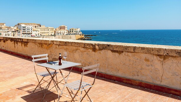 A terrace with a table and chairs overlooking a coastal cityscape. - Ortigia Terrazza Su Due Mari by Ortigia Apartm... (Syracuse)