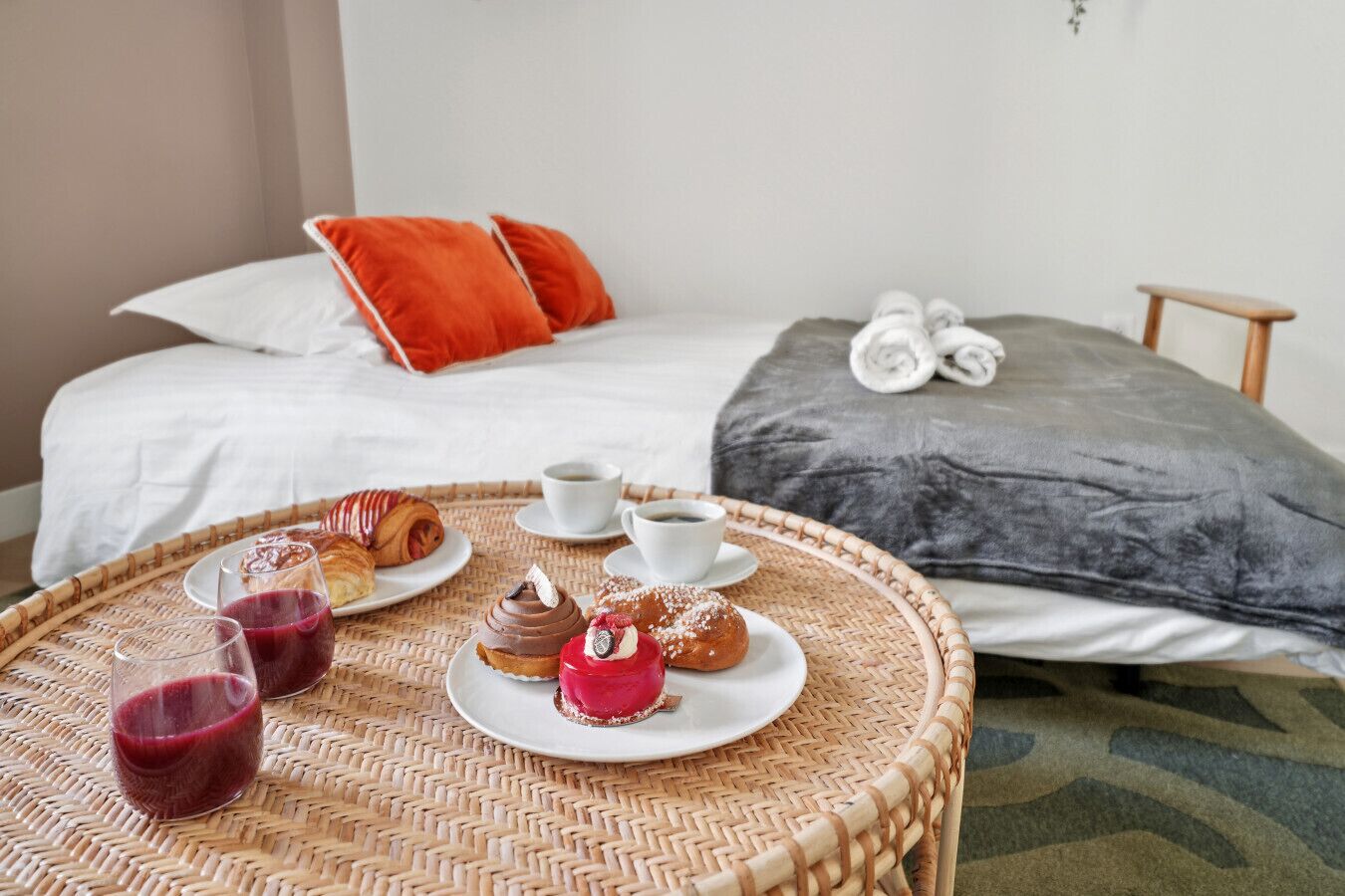 A neatly arranged breakfast table with pastries and drinks, a bed with a grey blanket and orange pillows, and a small wooden stool.