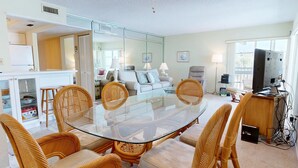 Un salon moderne avec une table à manger au plateau en verre, entourée de chaises en osier, d’un canapé blanc, d’un fauteuil gris et d’un téléviseur.