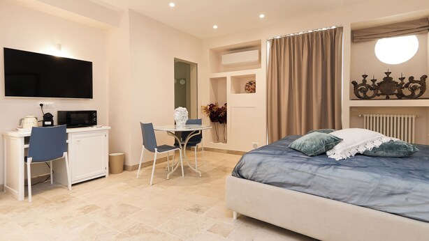 A hotel room with a bed, a desk with a chair, a television mounted on the wall, and a window with curtains. - Marina Homes Elegant designroom1 Ortigiapartments (Syracuse)