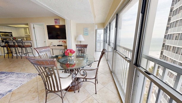 A dining area with a glass table and chairs, a TV, and a view of the sea. - Crescent Arms Vacation Rentals 703N Siesta Key Florida (Sarasota)