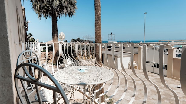 A balcony with ornate metal railings, a round table, and chairs, overlooking a beach with palm trees and a clear sky. - Marina Homes Deluxe Sea View 2 Ortigiapartments (Syracuse)