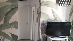 Olohuoneessa on valkoinen, litteänäyttöinen televisio, ovi, joka johtaa toiseen huoneeseen, ja seinä, jossa on trooppinen lehtikuvio.