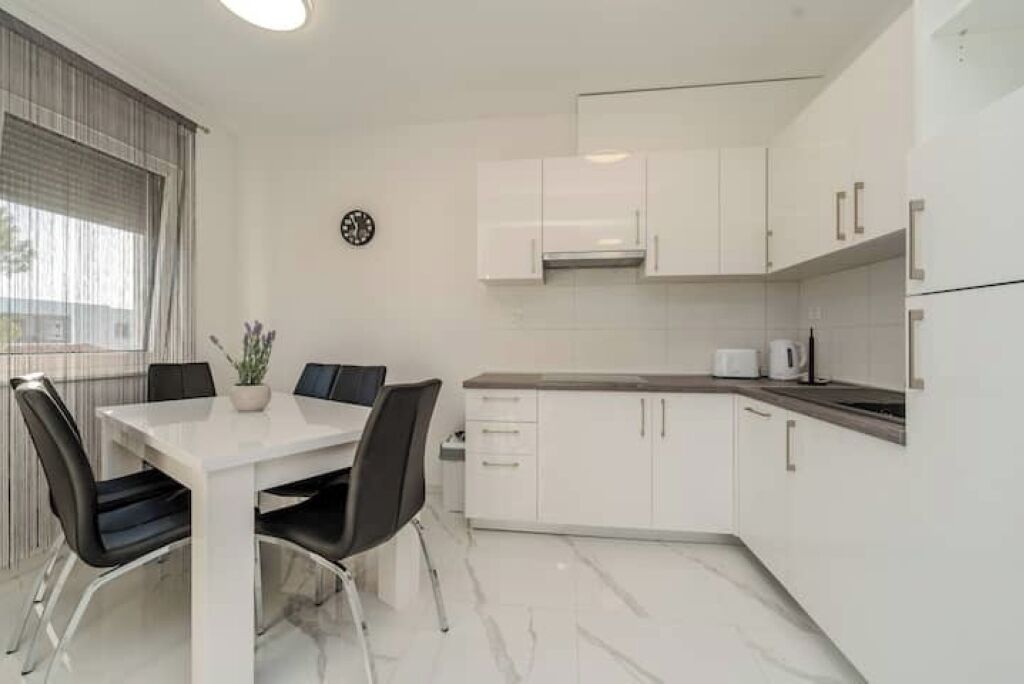 Een moderne keuken met witte kasten, een marmeren aanrechtblad en een eethoek met een witte tafel en zwarte stoelen.