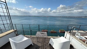 Terraza con dos sillas blancas y una mesa pequeña, con vista a un cuerpo de agua y una lejana vista de tierra y un bote.