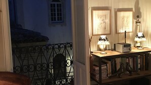 A room with a stone floor, a wooden chair, a small table with lamps, framed pictures, and a bookshelf. - Terrazza Cavalieri di Malta by Ortigiapartments (Syracuse)