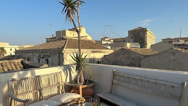 A rooftop terrace with a wicker sofa, a wooden bench, a table with snacks and drinks, and a potted palm tree. - Terrazza Cavalieri di Malta by Ortigiapartments (Syracuse)