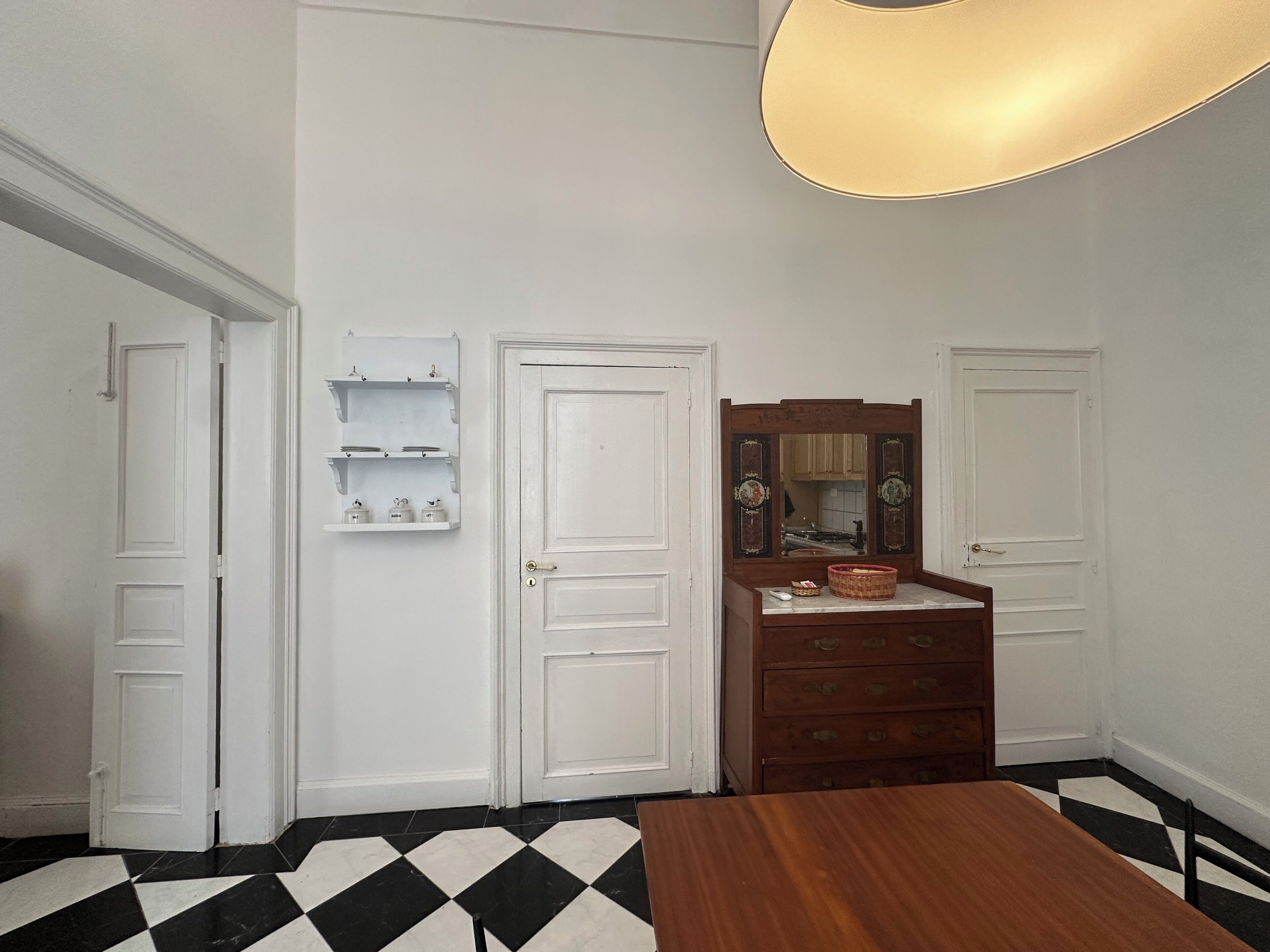 Quarto com piso xadrez, paredes brancas, cômoda de madeira, espelho e luminária pendente.