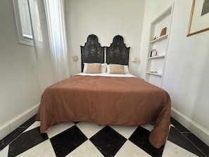 Ein ordentlich bezogenes Bett mit einer braunen Bettdecke, einem schwarz-weißen Schachfußboden und einem dunklen, mit floralen Mustern versehenen Kopfteil.